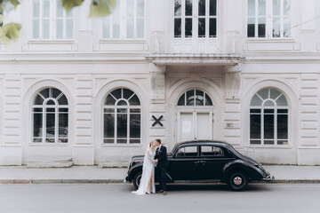 Wall Mural - Wedding of a beautiful, stylish couple, a bride in a white wedding dress and a groom in a black suit, tuxedo, in a black retro car.