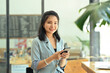 © saltdium - A portrait of a pretty Asian woman sitting in the office using a smartphone and looking at the camera with confidence, for business and technology concept.
