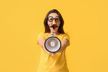Young Woman In Funny Disguise And With Megaphone On Yellow Background. April Fools' Day Celebration