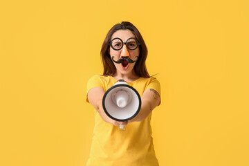 Young woman in funny disguise and with megaphone on yellow background. April fools' day celebration