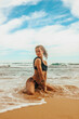 © NASTYA PALEHINA - vacation mood. young girl in black stylish swimsuit is sitting sexy in profile and smiling cute on sand  beach coast near wave of ocean  on blue sky background. travel lifestyle concept, free space