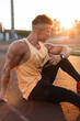 © alones - Handsome young stylish guy with a healthy athletic body in a yellow tank top, black leggings and sneakers sits on the street at sunset
