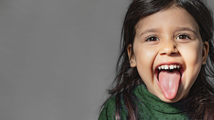 A pretty girl with dark hair and eyes sticks out her tongue on a gray background. Close up portrait of emotional preschooler in green longsleeve