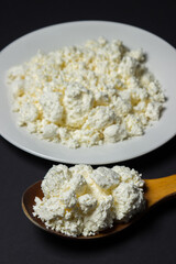 Wall Mural - A plate of cottage cheese on a black background. Healthy and tasty dairy product. Wooden spoon full of cottage cheese