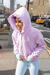 City lifestyle of young mixed race woman wearing pink hoodie with the word artist on it laughing with hood up