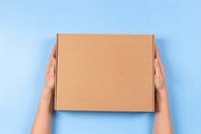 Top View To Female Hands Holding Brown Cardboard Box On Light Blue Background. Mockup Parcel Box. Packaging, Shopping, Delivery Concept