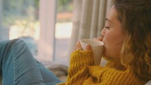 Young Woman In Cosy Warm Jumper Sitting In Chair At Home Looking Out Of Window With Hot Drink - Shot In Slow Motion