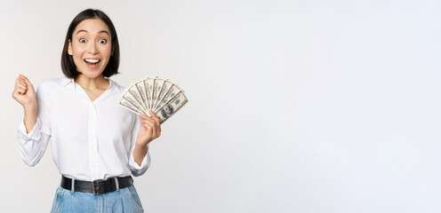 Enthusiastic young asian woman looking excited at camera, holding money dollars in hand, standing over white background