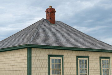 A vintage yellow cedar shake clapboard house with a grey shingled wooden hip roof. The building has a red brick chimney and multiple glass windows.  The sky in the background is blue with clouds.