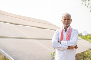 handheld shot of Smiling village farmer standing with crossed arms by looking at camera in front of solar panel - concept of modern agriculture, green energy and eco-friendly farming.