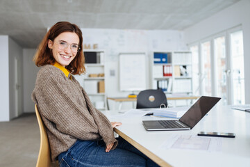 Wall Mural - young business woman with glasses sits at a desk with laptop and laughs into the camera