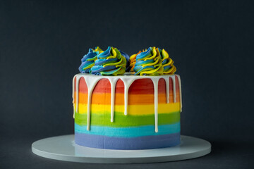 Canvas Print - Rainbow cake with whipped cream top on the dark grey background. Birthday cake with multicolored cream cheese frosting and white chocolate drips.