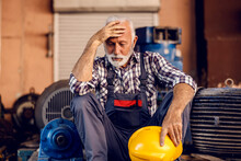 A Worry Senior Factory Worker Sits Next To The Machines And Holding His Head. 