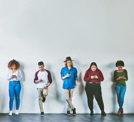 Poster - The always available generation. Studio shot of a group of young people standing against a wall and using their mobile phones.