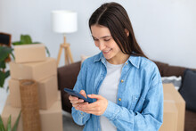 Moving To A New House, Rental Housing. Happy Young Caucasian Woman Using A Mobile Phone To Search And Order A Transportation Service And Movers To Move To A New Home