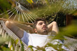 Jeune homme de 20 ans qui fait la sieste dans son jardin au soleil, allongé dans un hamac. Au premier plan, il y a des plantes.