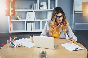 Wall Mural - Modern tech for modern designs. Shot of a young woman using a mobile phone and laptop in her design studio.