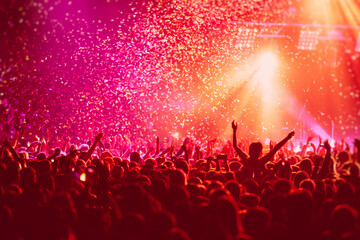 A crowded concert hall with scene stage in red lights, rock show performance, with people silhouette, colourful confetti explosion fired on dance floor air during a concert festival