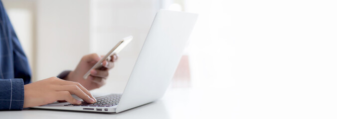 Closeup hand of young man working laptop computer and reading smartphone on internet online on desk at home, freelance male using phone with social media, business and communication concept.