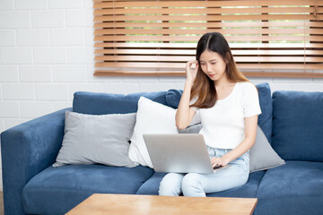 Wall Mural - Young asian business woman smile and work from home with laptop computer online to internet on sofa in living room, freelance girl using notebook on couch with comfort, new normal, lifestyle concept.