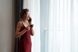 © DragonImages - Cheerful mature woman standing at window, drinking morning coffee and talking on phone with family member or colleague