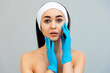 © _KUBE_ - Portrait of a young beautiful woman with a bandage on her head, leaning her gloved hands against her face. Gray background. The concept of professional cosmetology