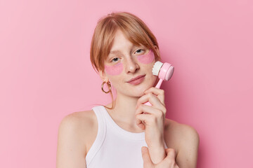 Serious redhaired millennial girl uses face massager applies hydrogel patches under eyes wears casual white t shirt isolated over pink studio background. Skin care and facial treatments concept