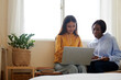 © DragonImages - Smiling multi-ethnic friends sitting on bench at window in dormitory and programming on laptop