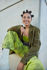 Wall Mural - Vertical shot of happy fashionable woman dressed in green costume looks away gladfully sits near concrete wall has good mood feels carefree spends free time outdoors. Youth and lifestyle concept