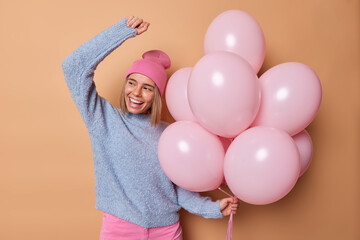 Wall Mural - Horizontal shot of positive European woman wears hat casual blue jumper dances carefree enjoys birthday celebration holds bunch of inflated balloons isolated over brown background. Festive occasion