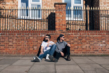 Two Men Sitting On Sidewalk In Front Of Brick Wall