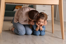 Scared Mother With Her Little Daughter Hiding Under Table In Living Room During Earthquake