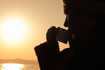 Young woman drinking Turkish coffee at dramatic sunset and watching distant sea on orange sunny day. Beautiful nature photography. Having fun time concept idea. Blank copy space or area for text.