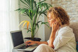 © simona - Side view of young modern adult people caucasian female using laptop sitting at home. Woman with blonde curly hair working on computer in smart job activity sitting on the sofa in indoor leisure