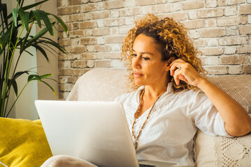 Wall Mural - Confident woman having relax leisure activity at home using laptop sitting on the sofa. Female people and computer technology smart working job. Young lady do video call in alternative office house