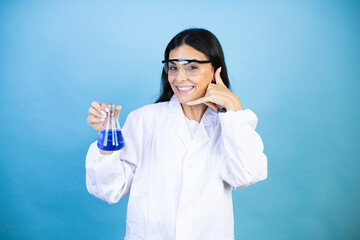 Wall Mural - Young brunette woman wearing scientist uniform holding test tube over isolated blue background smiling doing phone gesture with hand and fingers like talking on the telephone