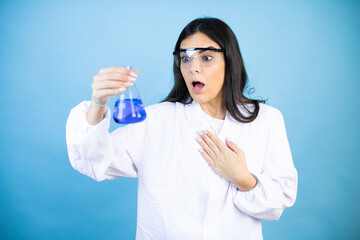 Wall Mural - Young brunette woman wearing scientist uniform holding test tube over isolated blue background afraid and shocked with surprise and amazed expression, fear and excited face.