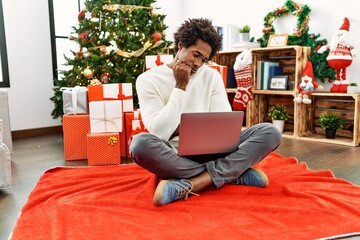 Poster - Young african american man using laptop sitting by christmas tree thinking looking tired and bored with depression problems with crossed arms.