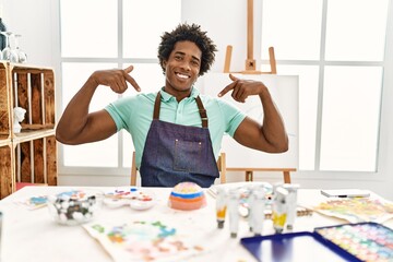 Wall Mural - Young african american man sitting on the table at art studio looking confident with smile on face, pointing oneself with fingers proud and happy.