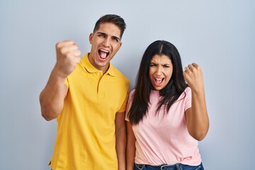 Poster - Young couple standing over isolated background angry and mad raising fist frustrated and furious while shouting with anger. rage and aggressive concept.