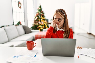 Poster - Young caucasian girl sitting on the table working using laptop by christmas tree smelling something stinky and disgusting, intolerable smell, holding breath with fingers on nose. bad smell