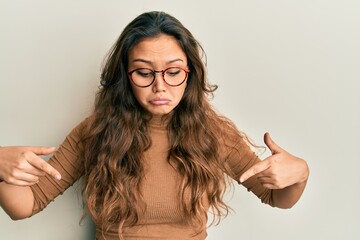 Wall Mural - Young hispanic girl wearing casual clothes and glasses pointing down looking sad and upset, indicating direction with fingers, unhappy and depressed.