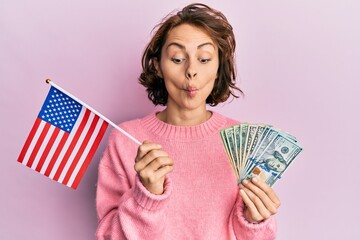 Poster - Young brunette woman holding united states flag and dollars making fish face with mouth and squinting eyes, crazy and comical.