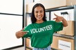 Young latin woman pointing with fingers to volunteer uniform at charity center