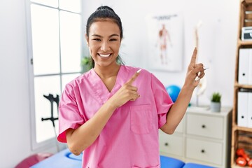 Sticker - Young hispanic woman working at pain recovery clinic smiling and looking at the camera pointing with two hands and fingers to the side.