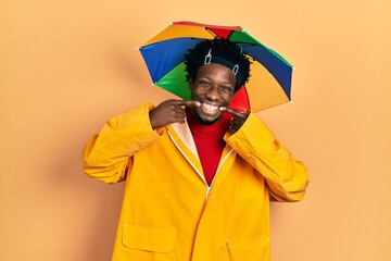 Sticker - Young african american man wearing yellow raincoat smiling cheerful showing and pointing with fingers teeth and mouth. dental health concept.