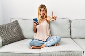 Wall Mural - Blonde beautiful young woman sitting on the sofa at home using smartphone pointing with finger up and angry expression, showing no gesture