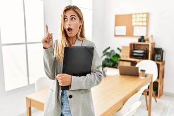 Canvas Print - Blonde business woman at the office amazed and surprised looking up and pointing with fingers and raised arms.