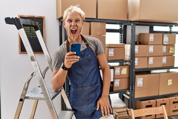 Canvas Print - Young blond man using smartphone working at storehouse angry and mad screaming frustrated and furious, shouting with anger. rage and aggressive concept.