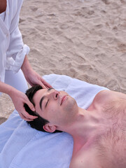 A relaxed young man lying on a massage table receiving a head massage on a beach in Valencia by a young chiromassage therapist.